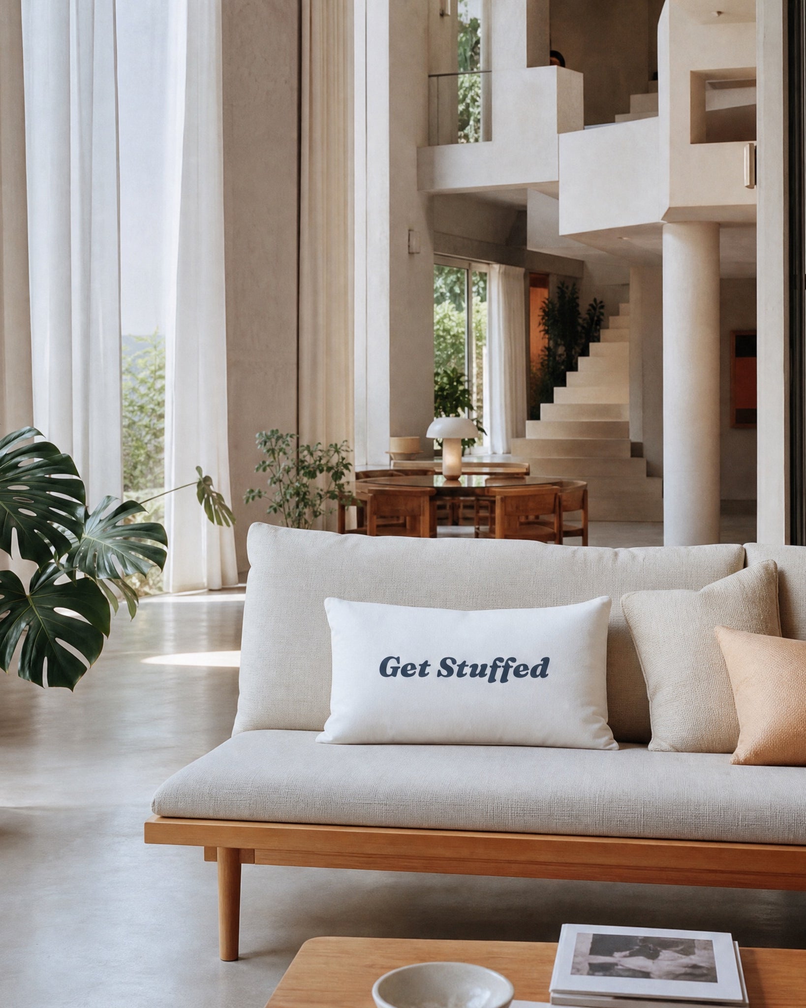 A white rectangular throw pillow with the text 'Get Stuffed' in navy blue script, styled on a light beige sofa in a modern, sunlit home interior.